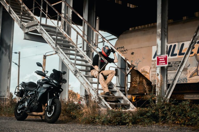 Motorcyclist takes a break under an industrial staircase beside a black motorcycle.