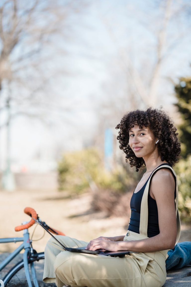 A curly-haired woman using a laptop in a sunny park, biking and working remotely.