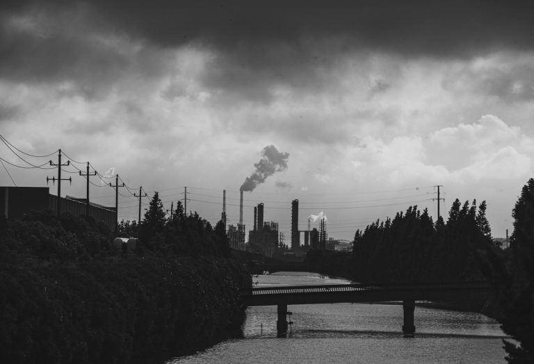 Black and white industrial scene with smokestacks emitting smoke over a river under cloudy skies.